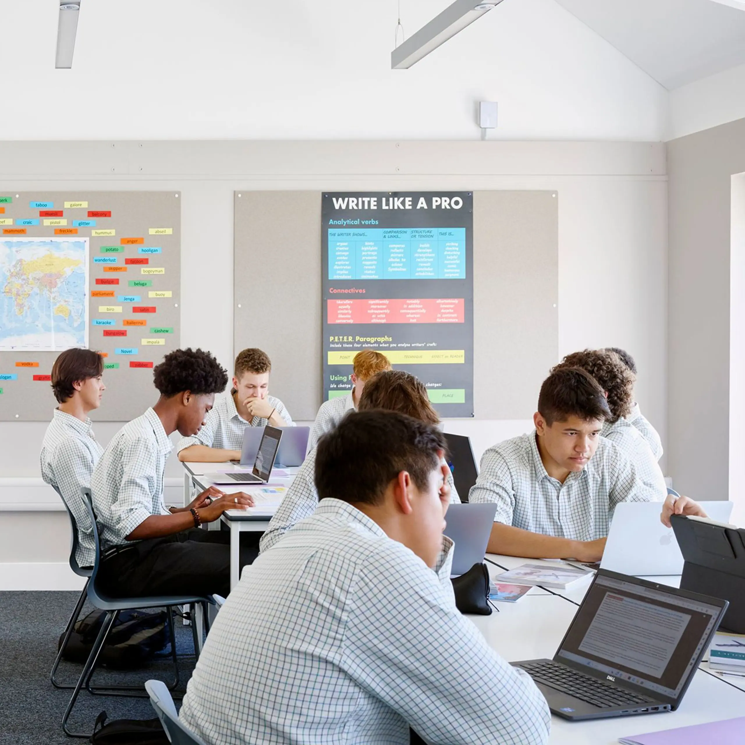Students working on laptops at desks inside a modern classroom with educational posters on the wall.