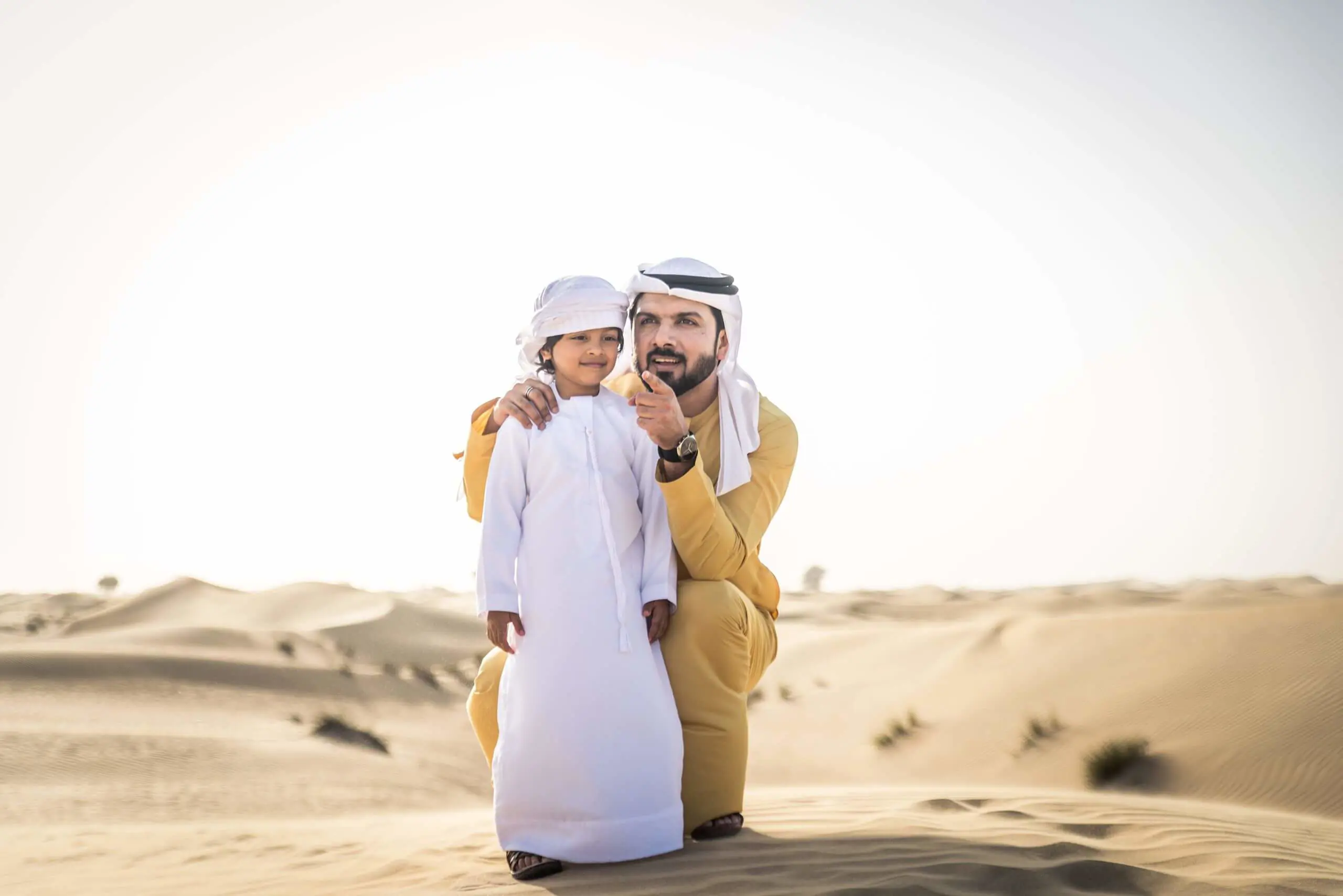 Father in traditional Emirati kandura standing with his young son outdoors in the desert dunes.