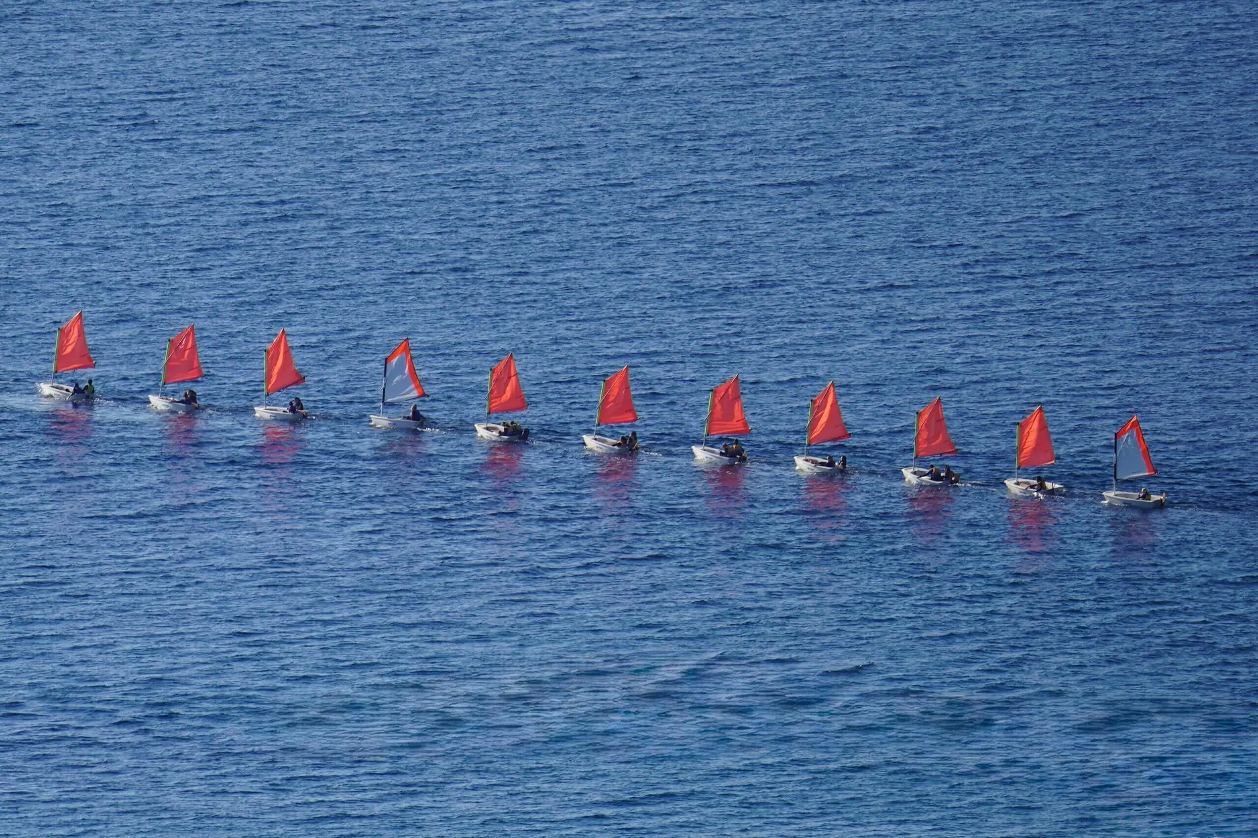 High aerial view of a line of red sailing boats navigating open blue water.