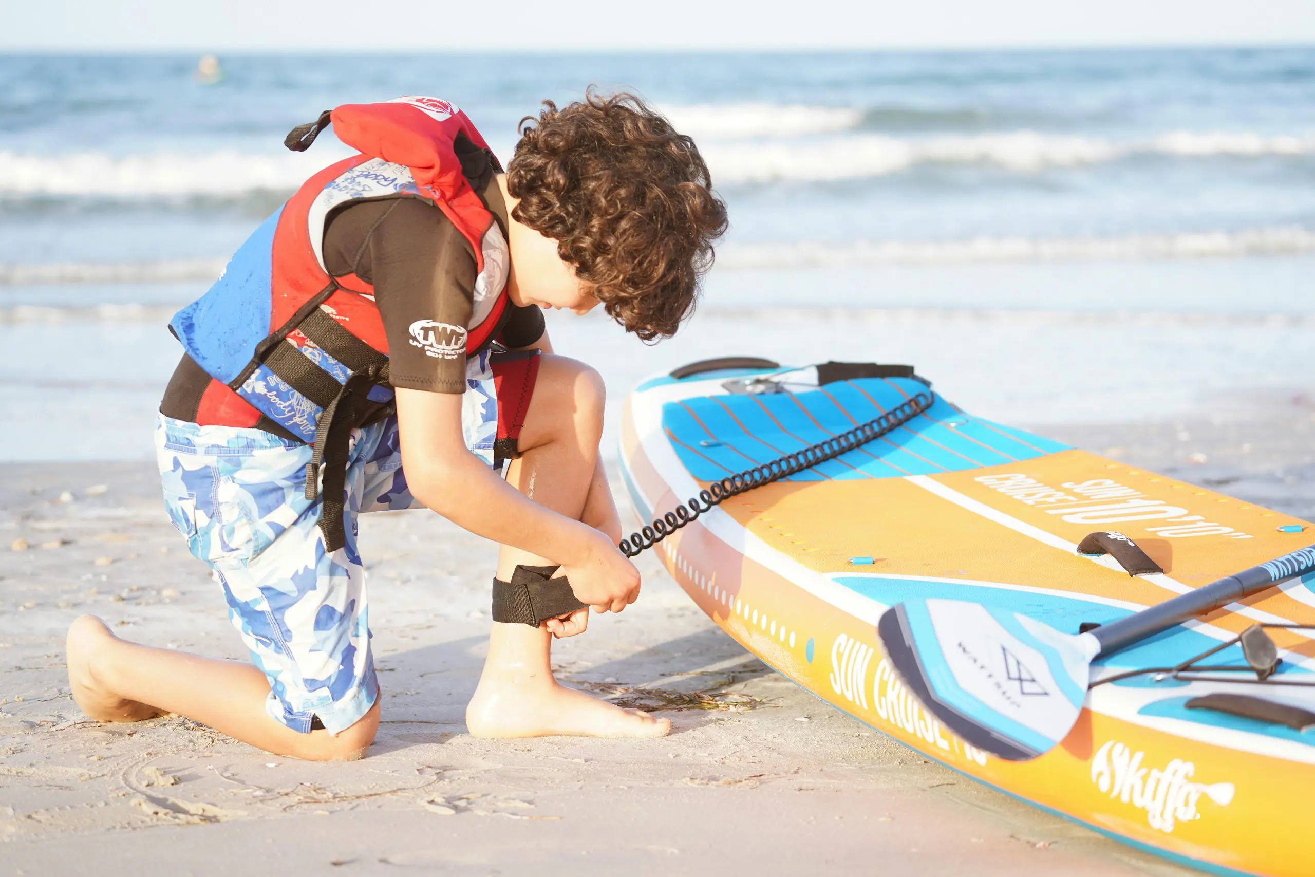 Young boy kneeling on a sandy beach securing a safety leash to his paddleboard.