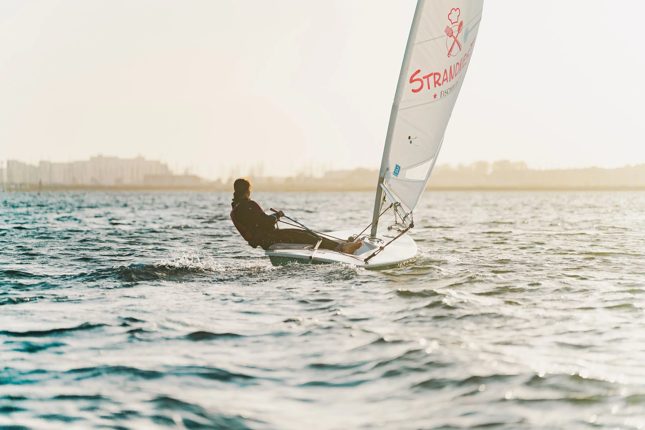 A student sailing a small sailboat on open coastal waters during sunrise.